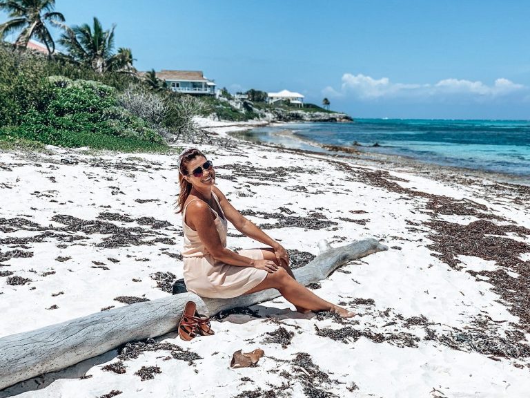 Ashley sitting on a large driftwood log, stuck in the white sand, looking out over the seaweed covered beach and blue waters of Cayman Kai Beach.