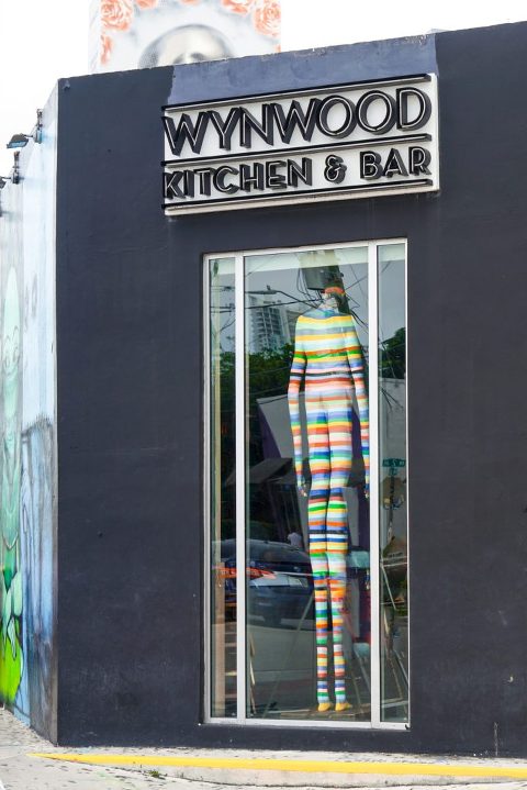 The outside of Wynwood Kitchen & Bar, featuring a large window display with a large colorful striped sculpture inside.