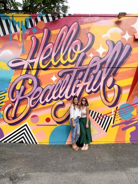Standing in front of a wall painted with the words "Hello Beautiful".