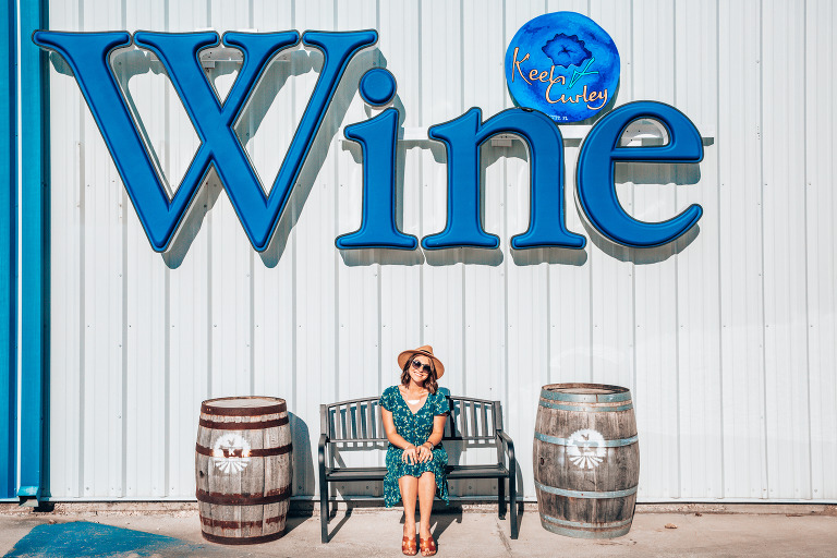 Dani sitting on a bench under the big blue "WINE" sign at Keel & Curley Winery