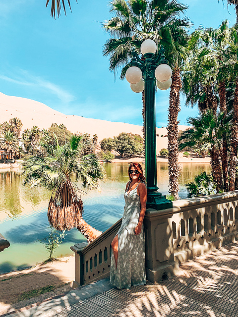 Ashley standing in front of the Huacachina Oasis. Blue green waters surrounded by desert dunes.